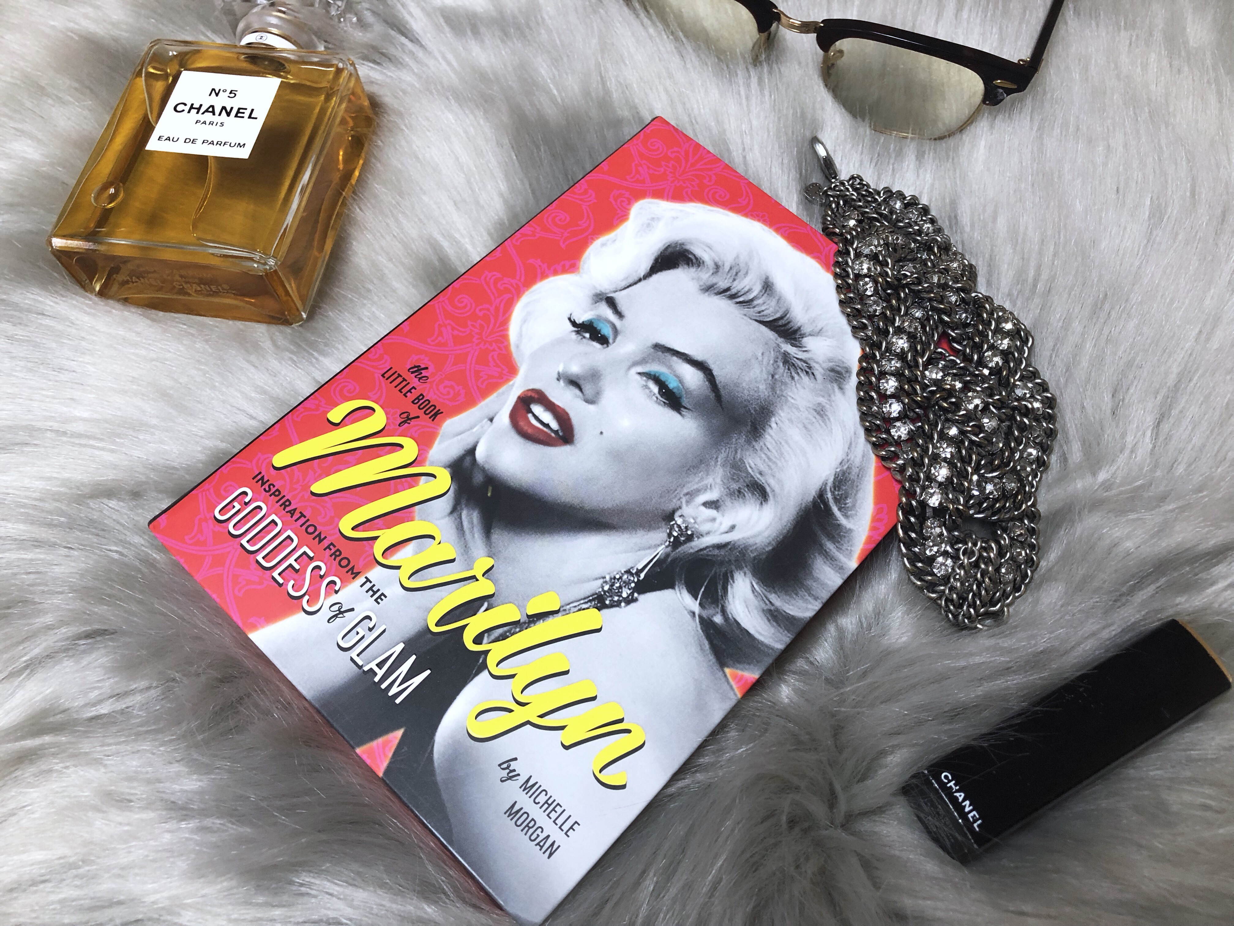 The little book of Marilyn, a little red book lies on a grey fur blanket with Chanel perfume and a diamond bracelet.